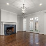 Empty living room with hardwood floors, a black tile fireplace, white walls, crown molding, a modern chandelier, and double French doors with white curtains leading outside.