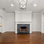 A bright, empty living room with dark hardwood floors, a central fireplace with a white mantel, white walls, French doors on the right, and an open view of a modern kitchen on the left.