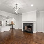 A spacious living room with hardwood floors, a black-tiled fireplace, white walls, and large windows. An open archway leads to a modern kitchen with white cabinets and stainless steel appliances.