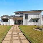 Modern white farmhouse with black roof, large windows, and a central entrance. A stone path leads through a well-kept lawn with landscaping and a white picket fence surrounds the yard under a clear blue sky.