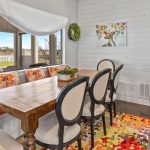 A cozy dining area with a wooden table, six chairs, floral cushions, and a colorful rug. A window seat lines one side, and a sideboard with a coffee maker and bottles stands against a white shiplap wall decorated with art and plates.