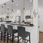 Modern kitchen with white cabinets, a long black countertop island with five dark gray upholstered bar stools, pendant lights above, stainless steel appliances, and an open view into a bright living space and bathroom.
