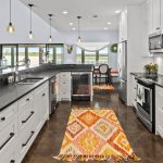 Modern kitchen with white cabinets, black countertops, stainless steel appliances, pendant lighting, large windows, and two colorful geometric rugs on a dark floor. Dining area visible in the background.