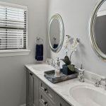 Modern bathroom with double sinks, silver-framed mirrors, gray cabinets, and a marble countertop. Window with blinds on the left, and a decorative tray with a plant and soap on the counter.