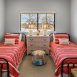 Two twin beds with red and white striped bedding are placed on either side of a white dresser with two lamps and decor, below a window with blinds. A colorful round ottoman sits between the beds. The walls are light gray.