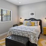 A cozy bedroom features a black wicker bed with a floral comforter, yellow and gray accent pillows, a yellow nightstand, wall-mounted lamps, a black bench, and a window with blinds letting in sunlight.