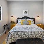 A neatly made bed with patterned bedding and yellow accent pillows is centered between two yellow nightstands. Wall lamps, artwork, and a window with blinds complete the cozy, modern bedroom decor.