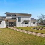 A modern white farmhouse with black trim, large windows, and metal roof, surrounded by a white picket fence and landscaped yard with stone path, trees, and garden beds under a clear blue sky.