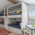 A cozy room with two sets of white built-in bunk beds, each with blue bedding and colorful pillows. A game table with two striped chairs sits near a window with wooden blinds. A red patterned rug is on the carpeted floor.