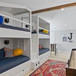 A bright room features two sets of white built-in bunk beds with navy bedding, colorful pillows, a red patterned rug, and a shuffleboard table. Artwork and a large “J” decorate the wall in the background.