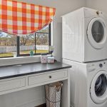 Bright laundry room with black countertop, white cabinets, a stacked washer and dryer, orange plaid window curtain, aprons hanging on the wall, and a window showing a white fence and trees outside.