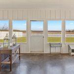 A bright sunroom with large windows and a glass door, showcasing views of a green lawn, white picket fence, and distant trees. Inside, there's wicker furniture, lanterns, and a side table on a concrete floor.