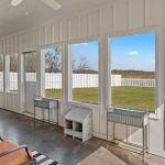 A bright sunroom with large windows, white paneled walls, and a view of a fenced backyard with grass and trees. There is patio furniture, metal planters, and decorative accents inside the room.