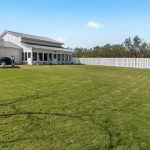 A modern white house with a metal roof sits beside a spacious, neatly mowed lawn, bordered by a white picket fence under a blue sky with a few clouds. Outdoor seating and a grill are visible on a patio.