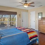 A cozy bedroom with a wooden headboard, blue bedding with a colorful striped blanket, large window with a view of trees, ceiling fan, dresser, framed pictures, and a guitar case against a beige wall.
