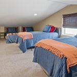 A cozy attic bedroom with four twin beds, each covered in blue bedding and orange throws. There are small windows, beige walls, and neutral carpeting, creating a warm and inviting atmosphere.