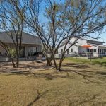 A fenced yard with two light-colored metal buildings, trees providing shade, a patio area with outdoor seating, and a large grassy lawn under a blue sky.