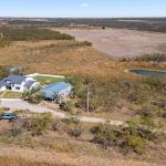 Aerial view of a rural property with a white house, a separate building, a circular driveway, a pond, scattered trees, and open fields under a clear sky.
