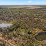 Aerial view of a vast, wooded landscape featuring two small ponds surrounded by sparse vegetation and trees, with open land and distant fields visible on the horizon under a blue sky.