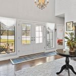 Bright entryway with large windows and a glass door, allowing sunlight to fill the space. Features a dark wood staircase, ornate cabinet, round table with plants, chandelier, white walls, and a view of a fenced yard outside.