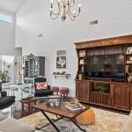 Bright living room with white shiplap walls, tall ceiling, large windows, and double doors. Features a wooden TV cabinet, sofas, armchairs with floral pillows, a rug, coffee table, and decorative accents.