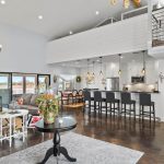 Open-concept living space with a modern kitchen, breakfast bar seating, gray sectional sofa, floral accent chairs, chandeliers, and a mezzanine above. Large windows provide natural light, and decor includes plants and animal art.