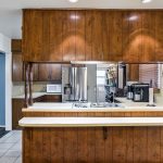 A kitchen with wooden cabinets, a double sink, and a beige countertop. Stainless steel refrigerator, microwave, coffee maker, and tiled floor are visible, with white walls and recessed ceiling lights.