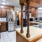 A kitchen with wooden cabinets, stainless steel refrigerator, microwave, gas stove, coffee machines, double sink, and white tile floor. A window with blinds lets in natural light above the sink area.