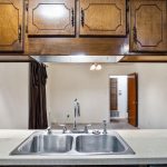Kitchen counter with double sink and chrome faucet, light-colored countertop, paper towel holder with a star-shaped top, and dark wood cabinets above. Open view to an adjacent room through a doorway.