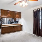 A kitchen with wooden cabinets and stainless steel appliances is separated from a carpeted dining area by a breakfast bar. The dining area has a window with dark brown curtains and a ceiling fan with a light fixture.