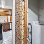 A kitchen with brown cabinets, white tile floor, and black appliances is adjacent to a small laundry area with a white washing machine and exposed insulation around the doorway.