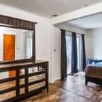 A bedroom with a large bed, blue and brown bedding, a dresser with a mirror, wood floors, and a sliding glass door with dark curtains letting in natural light.