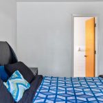 A bedroom with a bed covered in a blue geometric-patterned bedspread and pillows, next to a black headboard, with an open door revealing a white-tiled bathroom.