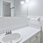 A clean, white bathroom with a sink, countertop, wall mirror, tile backsplash, light fixtures, towel rack, and a partially visible shower door. The decor is simple and minimalistic.