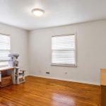 A bright, empty room with white walls, hardwood floors, two windows with blinds, a desk with a cat tree and supplies, and a large mirror on top of a wooden dresser. Ceiling light is on.