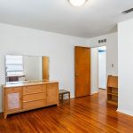 A simple bedroom with hardwood floors, a wooden dresser with a mirror, white walls, and two wooden doors—one to a closet and one to another room. A small shelf and a laundry basket are also visible.