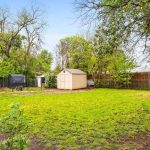 A grassy backyard with a wooden fence, scattered trees, two small animal enclosures, and a beige storage shed in the center. The sky is clear and blue.