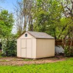 A beige wooden shed with a gray roof stands in a grassy backyard, surrounded by trees, a brown wooden fence, and wire fencing on a partly cloudy day.