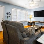 A cozy living room with gray sofas, a ceiling fan, and a TV mounted on a light blue wall. There's a table lamp by the window, a coffee table, hardwood floors, and a white door to the left.