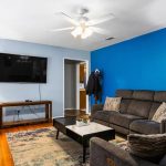A cozy living room with a blue accent wall, gray reclining sofas, a coffee table, a patterned rug, two tall lamps, a wall-mounted TV, and a ceiling fan with lights. A coat hangs on the wall near a doorway.