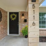A front porch with a black door decorated with a yellow wreath, brick walls, a house number "5116" on a wooden pillar, and a rain chain next to a flower bed and potted plants.