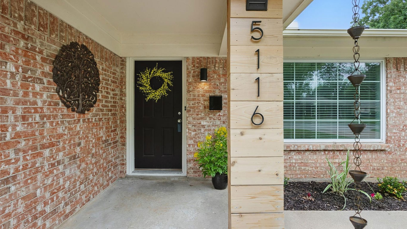 A front porch with a black door decorated with a yellow wreath, brick walls, a house number "5116" on a wooden pillar, and a rain chain next to a flower bed and potted plants.