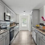Modern kitchen with light gray cabinets, granite countertops, stainless steel appliances, a sink with a potted orchid, tiled floor, and a window at the far end letting in natural light.