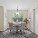 A bright dining area with a round table and four upholstered chairs, set on a blue and white rug. A chandelier hangs above, with a window, artwork, and a potted plant in the background. A door is visible on the left.