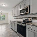 Modern kitchen with light gray cabinets, stainless steel appliances, a black and white tile backsplash, granite countertops, and gray tiled floors. A dining area with large windows is visible in the background.