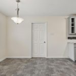 A bright, empty kitchen area with gray tile flooring, light gray cabinets, a white door in the center, and a large window letting in natural light. A hanging light fixture is above the eating area.