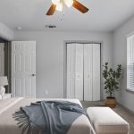 A bright bedroom with a bed, blue and beige bedding, a white bench, a potted plant, a lamp, wall art, two closed doors, and large windows letting in natural light.