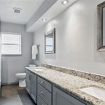 A modern bathroom with gray cabinets, a long granite countertop with two sinks, a large mirror, gray tile floor, white walls, and a window above the toilet at the far end. Towels hang on the wall near the sinks.