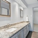 A modern bathroom with a double-sink granite vanity, two framed mirrors, light gray cabinets, recessed lighting, and a towel ring, leading to a carpeted bedroom with a window.