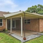A covered patio attached to a brick house with a rain chain, concrete floor, and gravel border in a grassy backyard. Trees and a wooden fence are visible in the background.